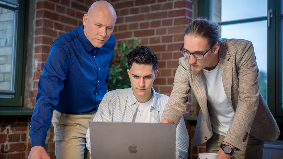 Drei Männer stehen und sitzen vor einem Laptop in einem modernen Büro mit Backsteinwänden. Zwei von ihnen lehnen sich engagiert über den Bildschirm, während der sitzende Mann konzentriert arbeitet. Die Szene vermittelt Teamarbeit und Mitarbeitermotivation.